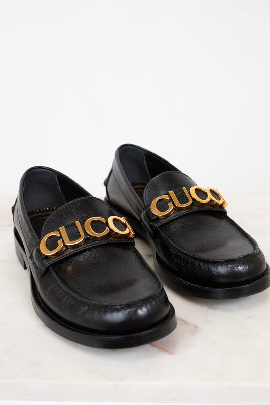 Black loafers with gold 'Gucci' logo on a light background