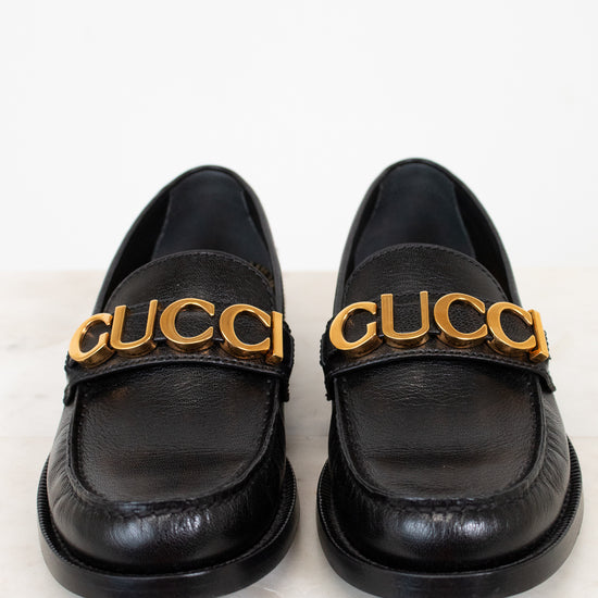 Pair of black loafers with gold 'Gucci' logo on a white background