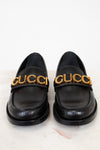 Pair of black loafers with gold 'Gucci' logo on a white background