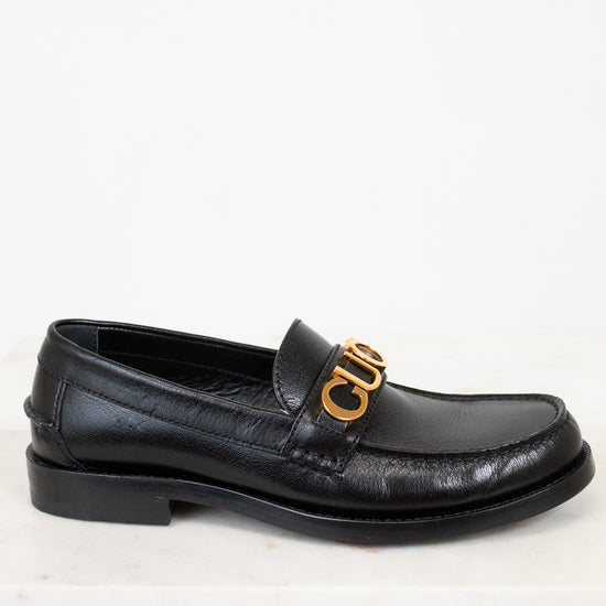 Black loafer shoe with gold logo on a white background