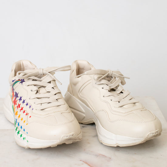A pair of chunky low-top sneakers in a light cream or off-white color, featuring a colorful rainbow star pattern on the side of the shoe, resting on a light marble surface against a white wall.