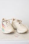 A pair of chunky low-top sneakers in a light cream or off-white color, featuring a colorful rainbow star pattern on the side of the shoe, resting on a light marble surface against a white wall.