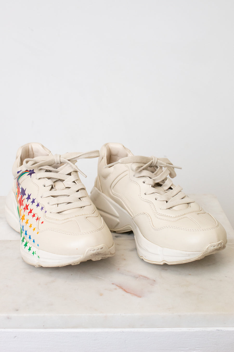 A pair of chunky low-top sneakers in a light cream or off-white color, featuring a colorful rainbow star pattern on the side of the shoe, resting on a light marble surface against a white wall.