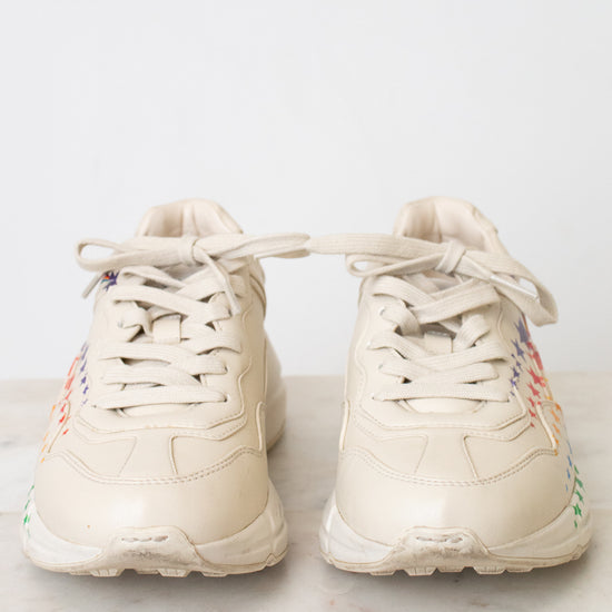 A pair of chunky low-top sneakers in a light cream or off-white color, featuring multicolor details on the sides, displayed on a light marble surface against a white background.