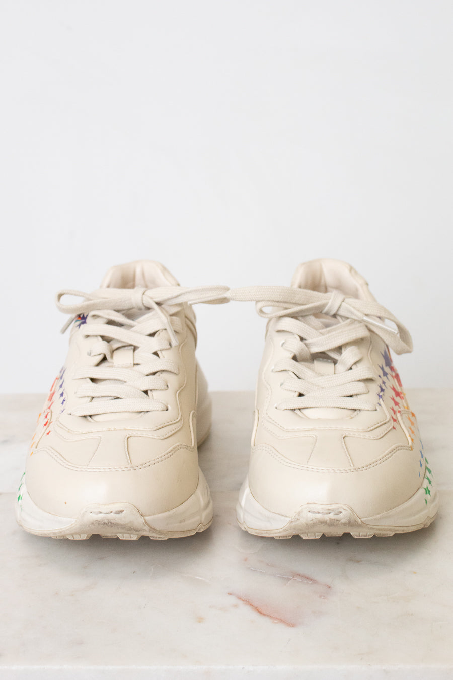 A pair of chunky low-top sneakers in a light cream or off-white color, featuring multicolor details on the sides, displayed on a light marble surface against a white background.