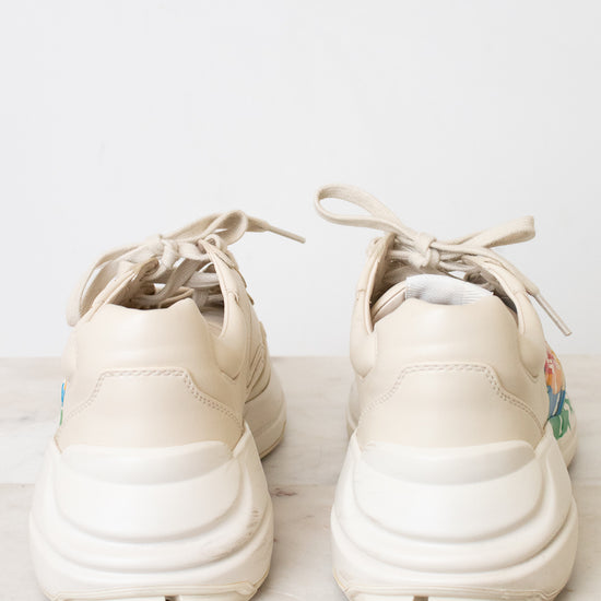 A pair of chunky platform sneakers in a light cream or off-white color, shown from the back on a light marble surface against a white background.