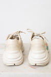 A pair of chunky platform sneakers in a light cream or off-white color, shown from the back on a light marble surface against a white background.