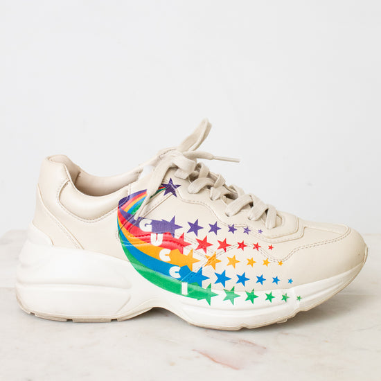 A chunky low-top sneaker in a light cream or off-white color, featuring a colorful rainbow star pattern on the side of the shoe, resting on a light marble surface against a white wall.