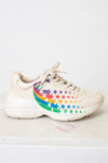 A chunky low-top sneaker in a light cream or off-white color, featuring a colorful rainbow star pattern on the side of the shoe, resting on a light marble surface against a white wall.