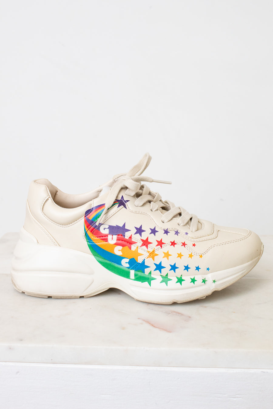 A chunky low-top sneaker in a light cream or off-white color, featuring a colorful rainbow star pattern on the side of the shoe, resting on a light marble surface against a white wall.