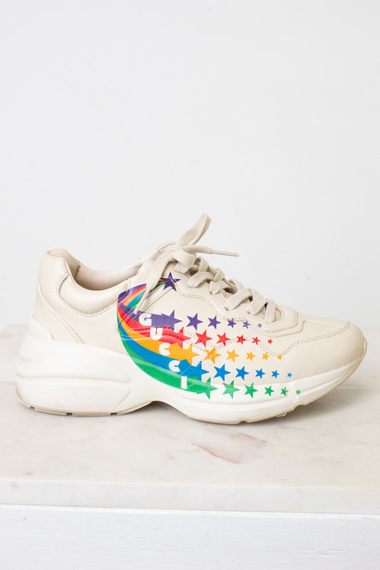 A chunky low-top sneaker in a light cream or off-white color, featuring a colorful rainbow star pattern on the side of the shoe, resting on a light marble surface against a white wall.