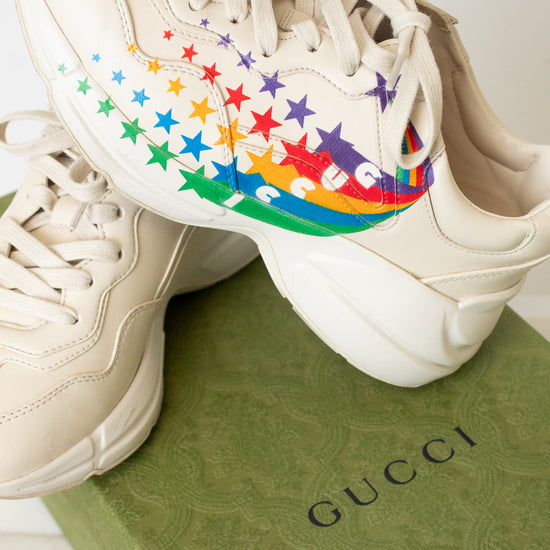 A pair of chunky low-top sneakers in a light cream or off-white color, featuring a colorful rainbow star pattern on the side of the shoe, sitting on top of a green "Gucci" box. 
