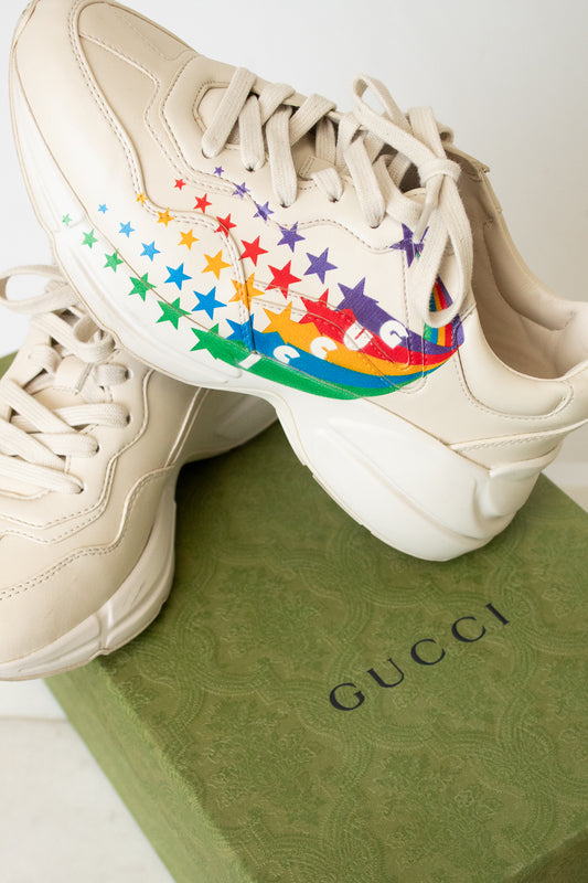 A pair of chunky low-top sneakers in a light cream or off-white color, featuring a colorful rainbow star pattern on the side of the shoe, sitting on top of a green "Gucci" box. 