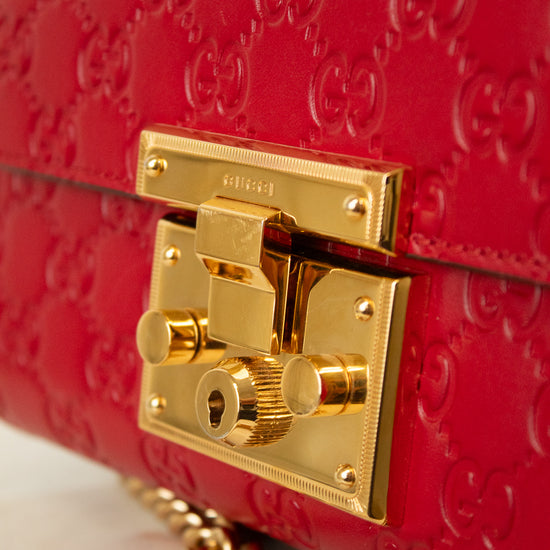 Close-up of a red Gucci shoulder bag with embossed GG monogram pattern and a prominent gold-tone lock closure. A gold chain strap is partially visible at the bottom left.