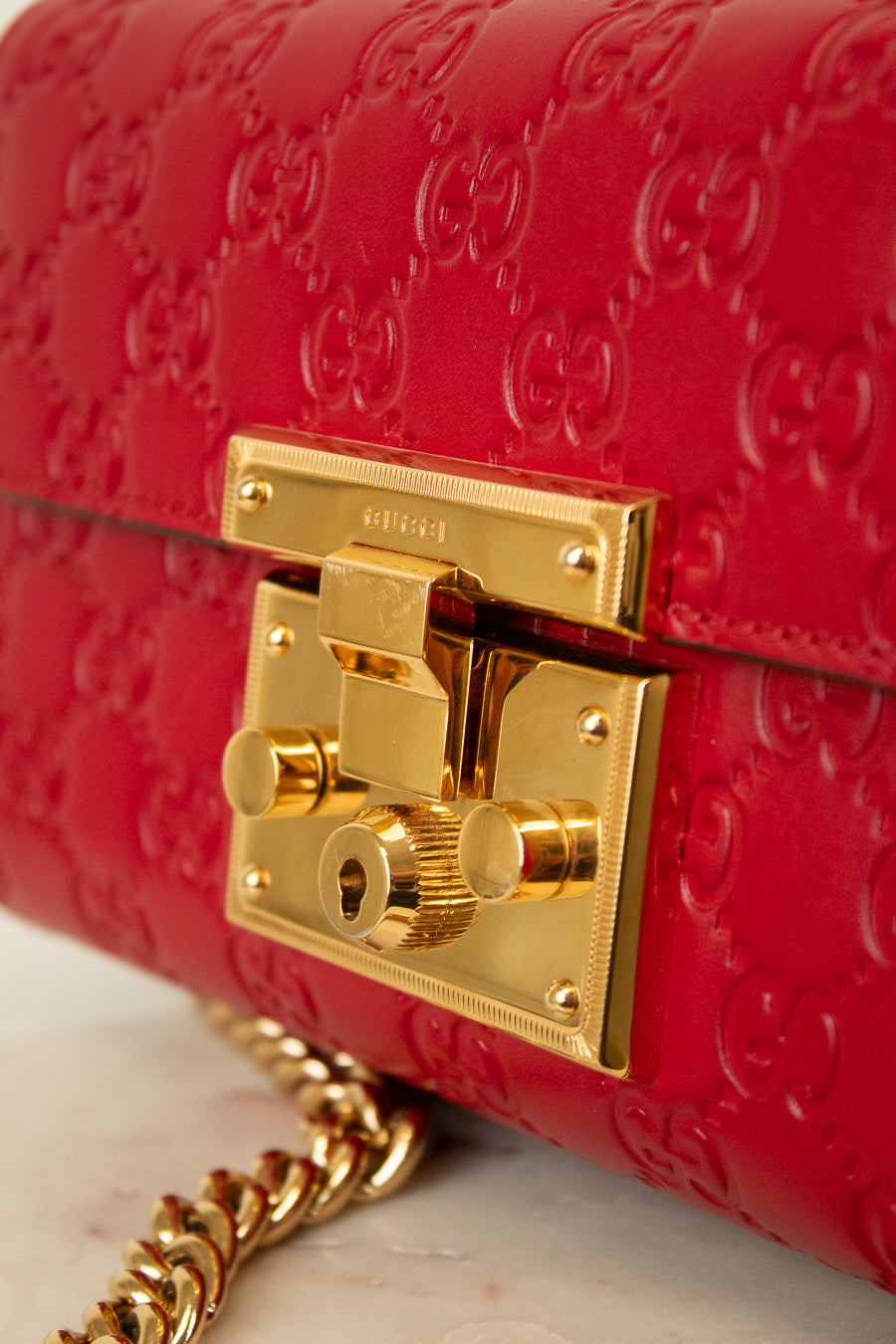 Close-up of a red Gucci shoulder bag with embossed GG monogram pattern and a prominent gold-tone lock closure. A gold chain strap is partially visible at the bottom left.