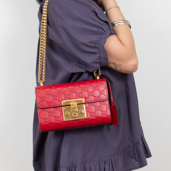 A woman wearing a purple ruffled dress carries a red quilted shoulder bag with a gold chain strap and gold clasp against a light gray background.