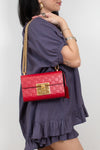 A woman wearing a purple ruffled dress carries a red quilted shoulder bag with a gold chain strap and gold clasp against a light gray background.