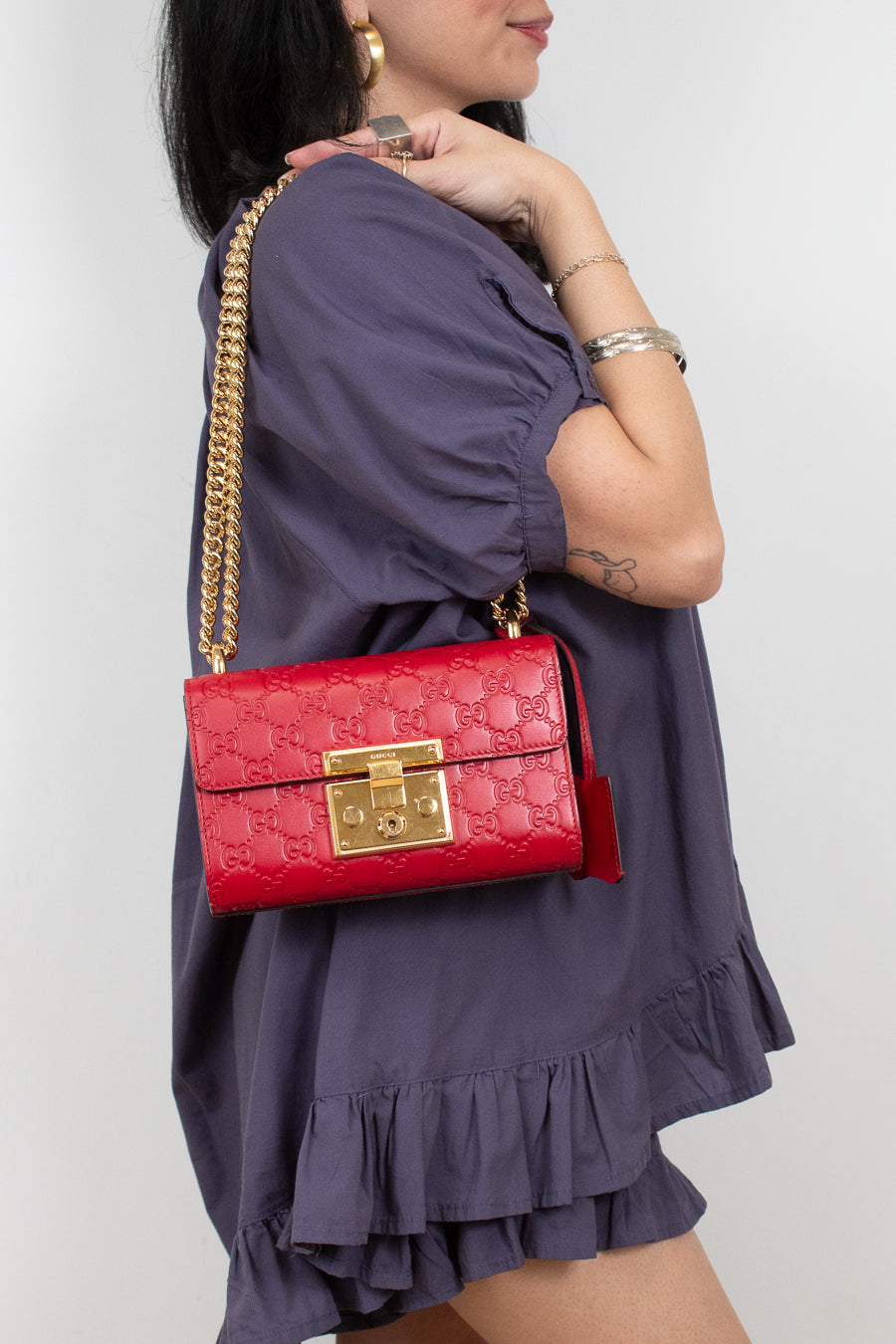 A woman wearing a purple ruffled dress carries a red quilted shoulder bag with a gold chain strap and gold clasp against a light gray background.