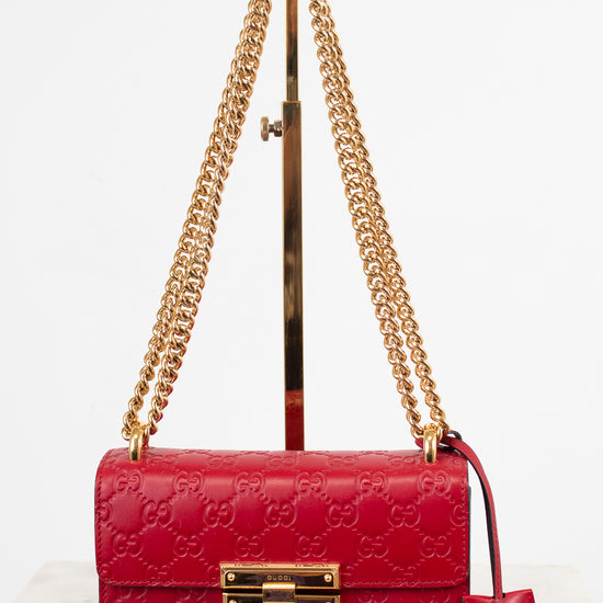 A red Gucci shoulder bag with embossed GG monogram pattern, gold-tone hardware, and a gold chain strap, displayed on a stand against a white background.