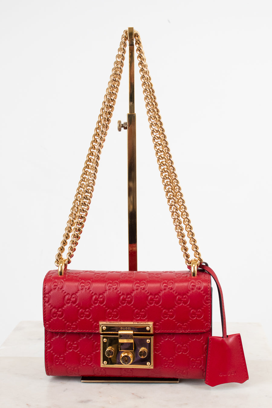A red Gucci shoulder bag with embossed GG monogram pattern, gold-tone hardware, and a gold chain strap, displayed on a stand against a white background.