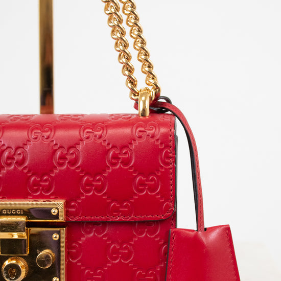 A close-up of a red Gucci shoulder bag featuring the embossed GG monogram, a gold-tone chain strap, and a matching red leather clochette with the Gucci logo, set against a clean white background.