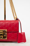 A close-up of a red Gucci shoulder bag featuring the embossed GG monogram, a gold-tone chain strap, and a matching red leather clochette with the Gucci logo, set against a clean white background.