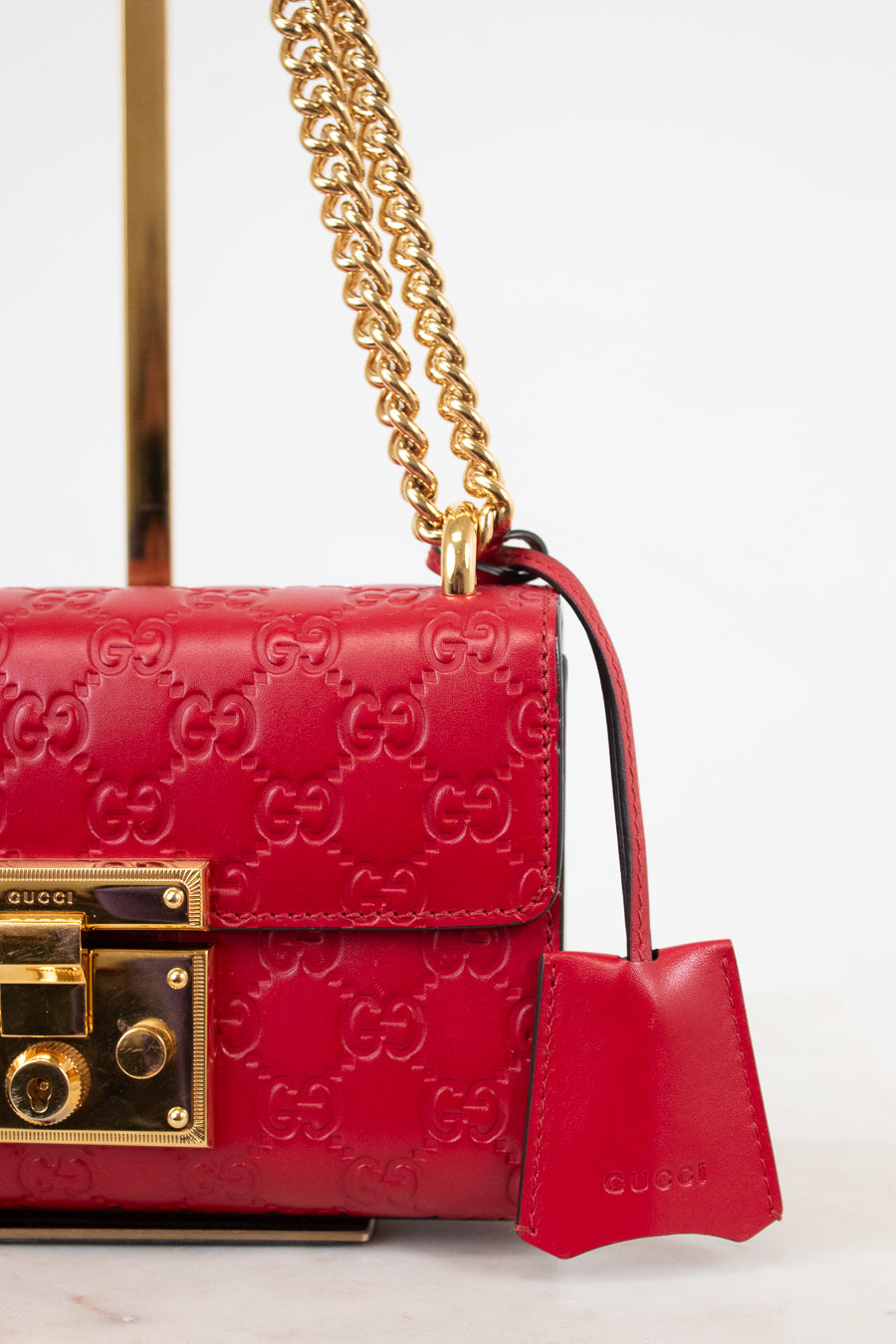 A close-up of a red Gucci shoulder bag featuring the embossed GG monogram, a gold-tone chain strap, and a matching red leather clochette with the Gucci logo, set against a clean white background.