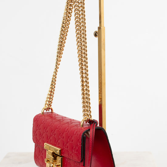 A red embossed leather shoulder bag with gold-tone chain straps and a gold clasp, displayed on a gold stand against a white background.
