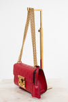 A red embossed leather shoulder bag with gold-tone chain straps and a gold clasp, displayed on a gold stand against a white background.