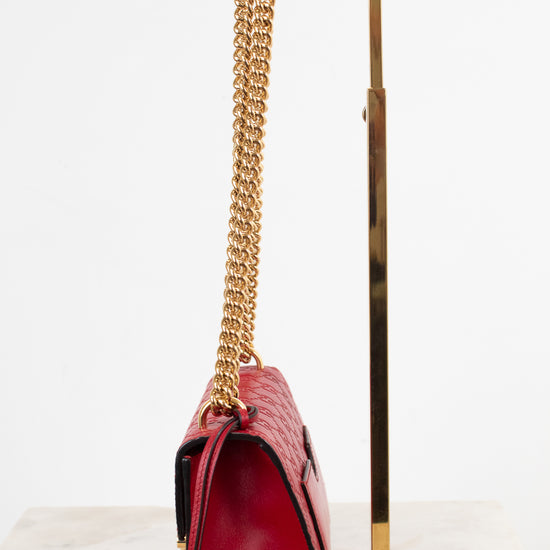A red crocodile-embossed leather handbag with a gold chain strap, displayed on a gold metal stand against a white background.