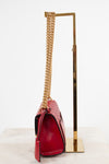 A red crocodile-embossed leather handbag with a gold chain strap, displayed on a gold metal stand against a white background.
