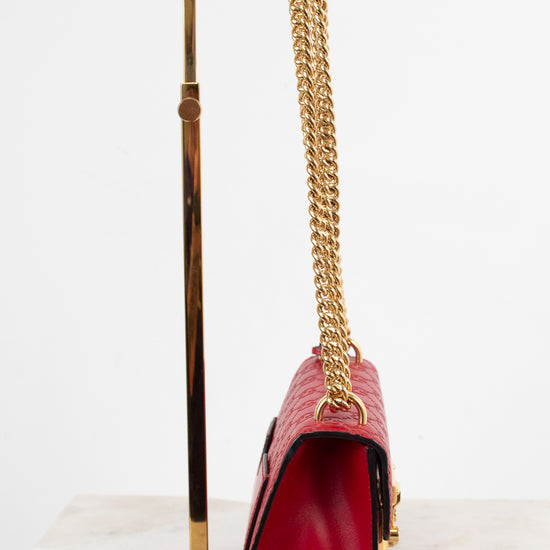 A red leather shoulder bag with a gold chain strap and gold hardware, displayed on a gold metal stand against a white background.