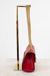 A red leather shoulder bag with a gold chain strap and gold hardware, displayed on a gold metal stand against a white background.