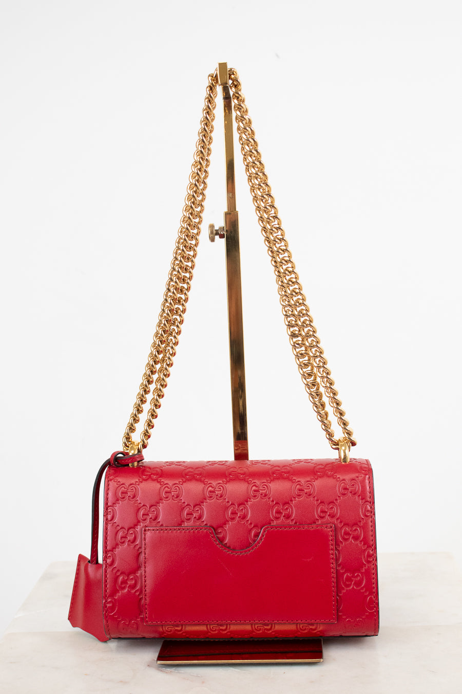 A red Gucci shoulder bag with embossed GG monogram pattern, gold chain strap, and gold-tone hardware, displayed on a stand against a white background.