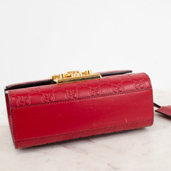 A red leather clutch bag with embossed GG monogram pattern and a gold-tone clasp, resting on a light marble surface against a white background.