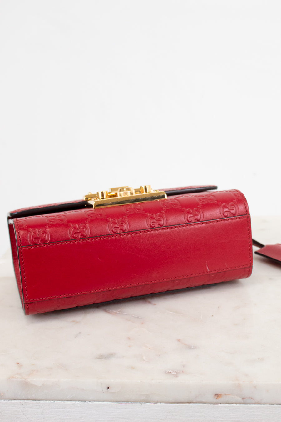 A red leather clutch bag with embossed GG monogram pattern and a gold-tone clasp, resting on a light marble surface against a white background.