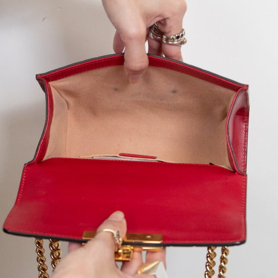 A close-up of a red leather handbag with gold chain straps, held open by two hands wearing rings and bracelets against a plain white background.