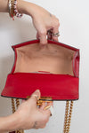 A close-up of a red leather handbag with gold chain straps, held open by two hands wearing rings and bracelets against a plain white background.