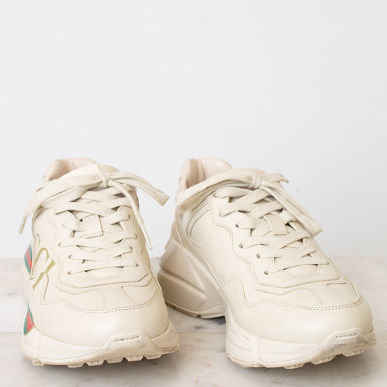 A pair of off-white Gucci chunky sneakers with gold lettering, the signature green and red web stripe, and a cream sole, displayed on a light marble surface against a white background.