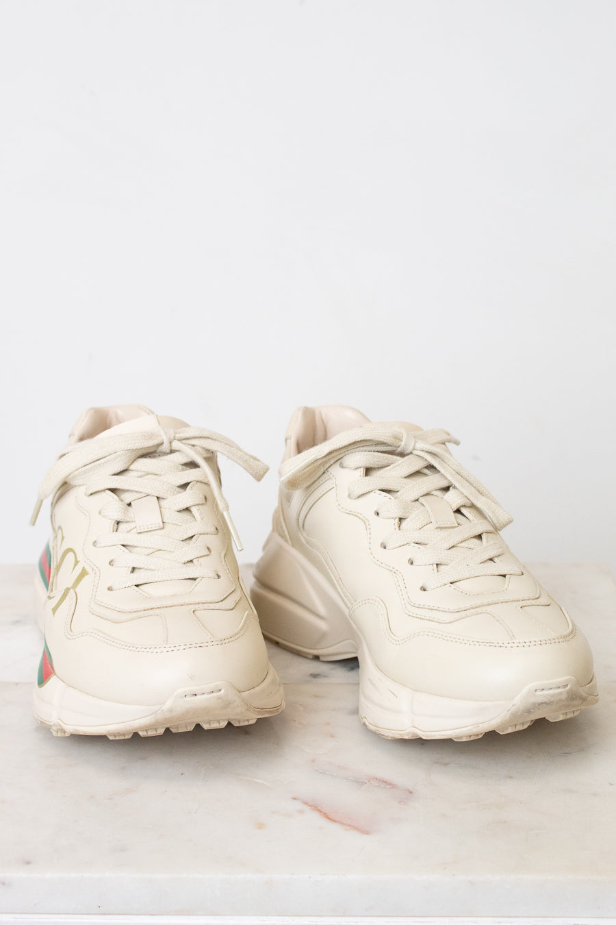 A pair of off-white Gucci chunky sneakers with gold lettering, the signature green and red web stripe, and a cream sole, displayed on a light marble surface against a white background.