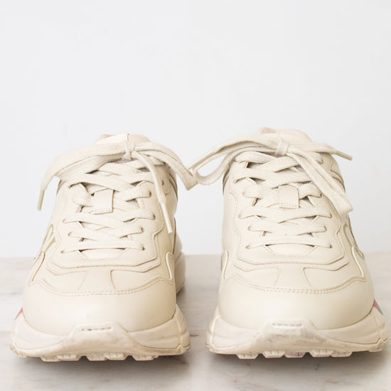 A pair of chunky low-top sneakers in a light cream or off-white color, shown from the front on a light marble surface against a white background.