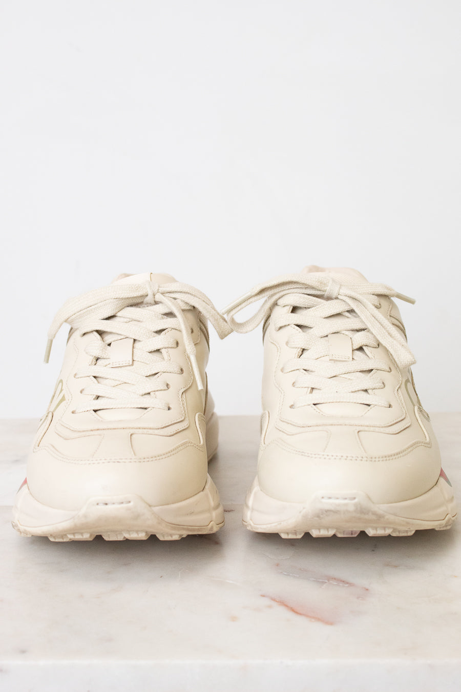 A pair of chunky low-top sneakers in a light cream or off-white color, shown from the front on a light marble surface against a white background.