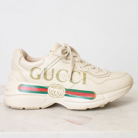 A single Gucci low-top sneaker in off-white leather with gold 'GUCCI' lettering on the side, a green and red web stripe, and a gold interlocking GG logo, shown against a light background.