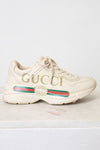 A single Gucci low-top sneaker in off-white leather with gold 'GUCCI' lettering on the side, a green and red web stripe, and a gold interlocking GG logo, shown against a light background.