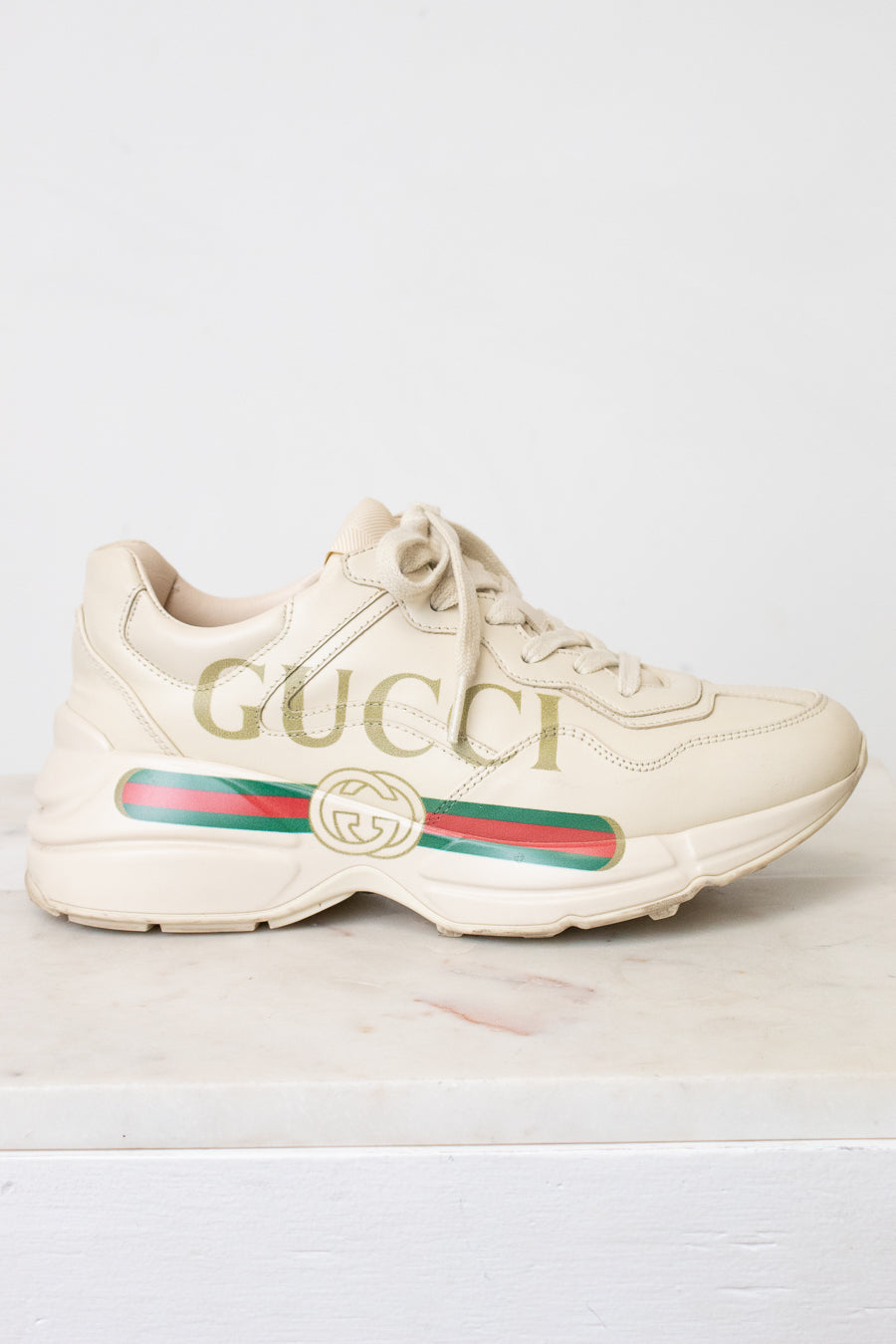 A single Gucci low-top sneaker in off-white leather with gold 'GUCCI' lettering on the side, a green and red web stripe, and a gold interlocking GG logo, shown against a light background.