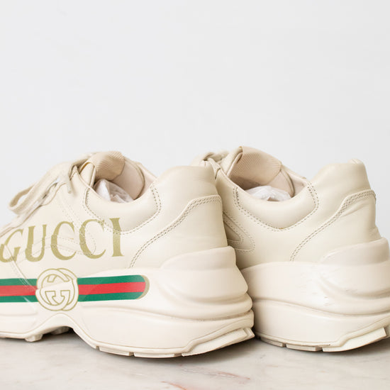 A pair of off-white Gucci chunky sneakers with gold lettering, the signature green and red web stripe, and a cream sole, displayed on a light marble surface against a white background.