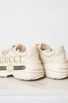 A pair of off-white Gucci chunky sneakers with gold lettering, the signature green and red web stripe, and a cream sole, displayed on a light marble surface against a white background.