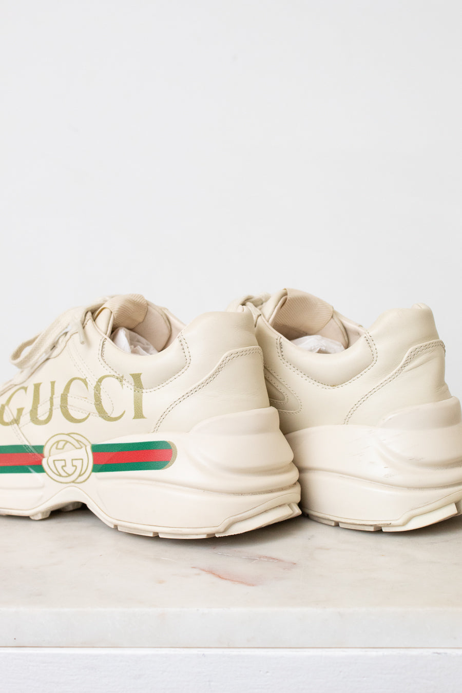 A pair of off-white Gucci chunky sneakers with gold lettering, the signature green and red web stripe, and a cream sole, displayed on a light marble surface against a white background.