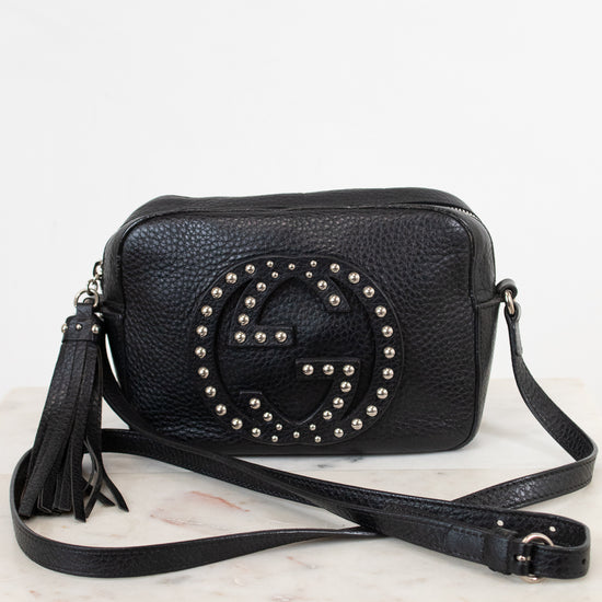 A black pebbled leather crossbody bag with a large studded interlocking G logo on the front, resting on a white marble surface against a white background.