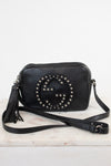 A black pebbled leather crossbody bag with a large studded interlocking G logo on the front, resting on a white marble surface against a white background.